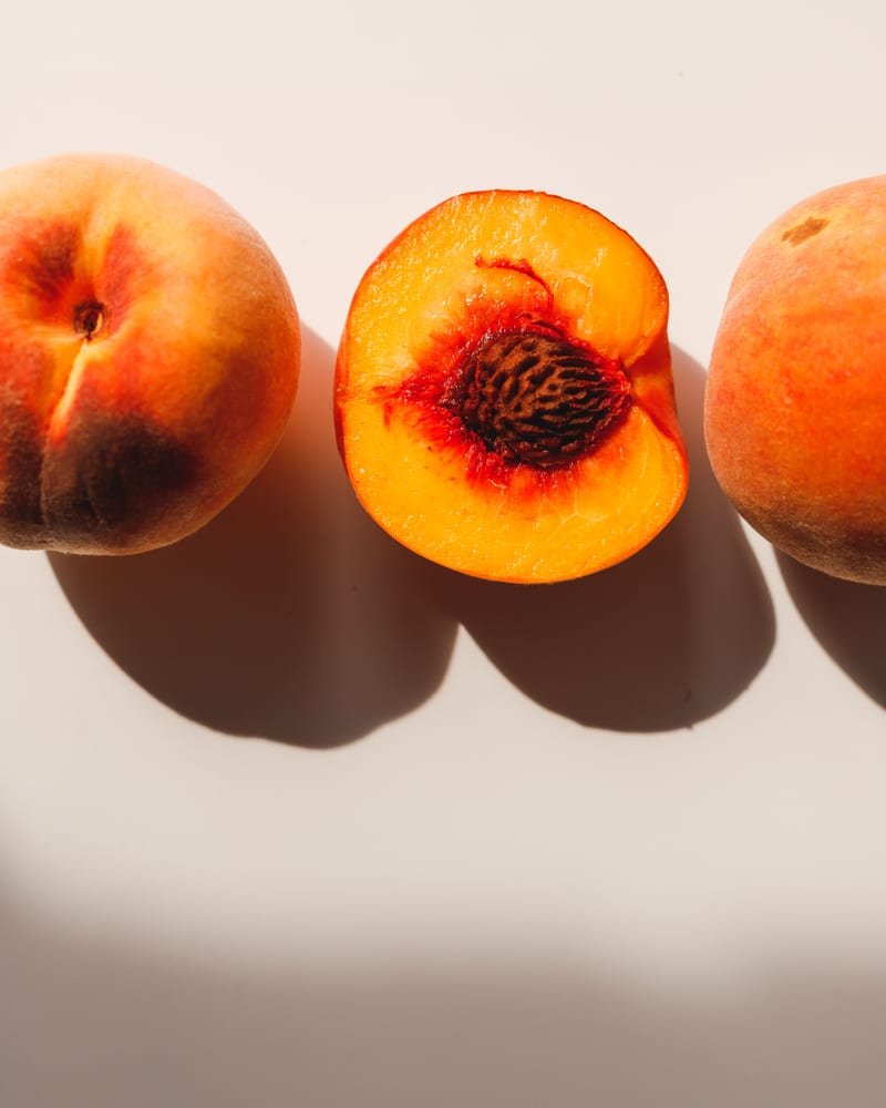 Three ripe peaches on a pale background, the middle one halved to show the stone.